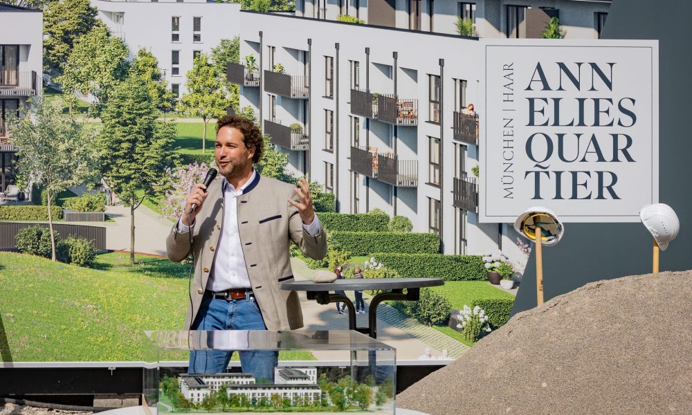 Bürgermeister Dr. Andreas Bukowski beim Spatenstich präsentiert Annelies Quartier mit Mikrofon und Werbeplane im Hintergrund